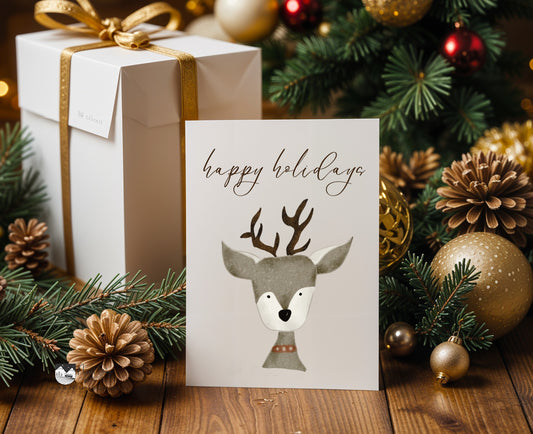 Christmas card with deer illustration and 'happy holidays' text, placed in front of a decorated Christmas tree with ornaments.