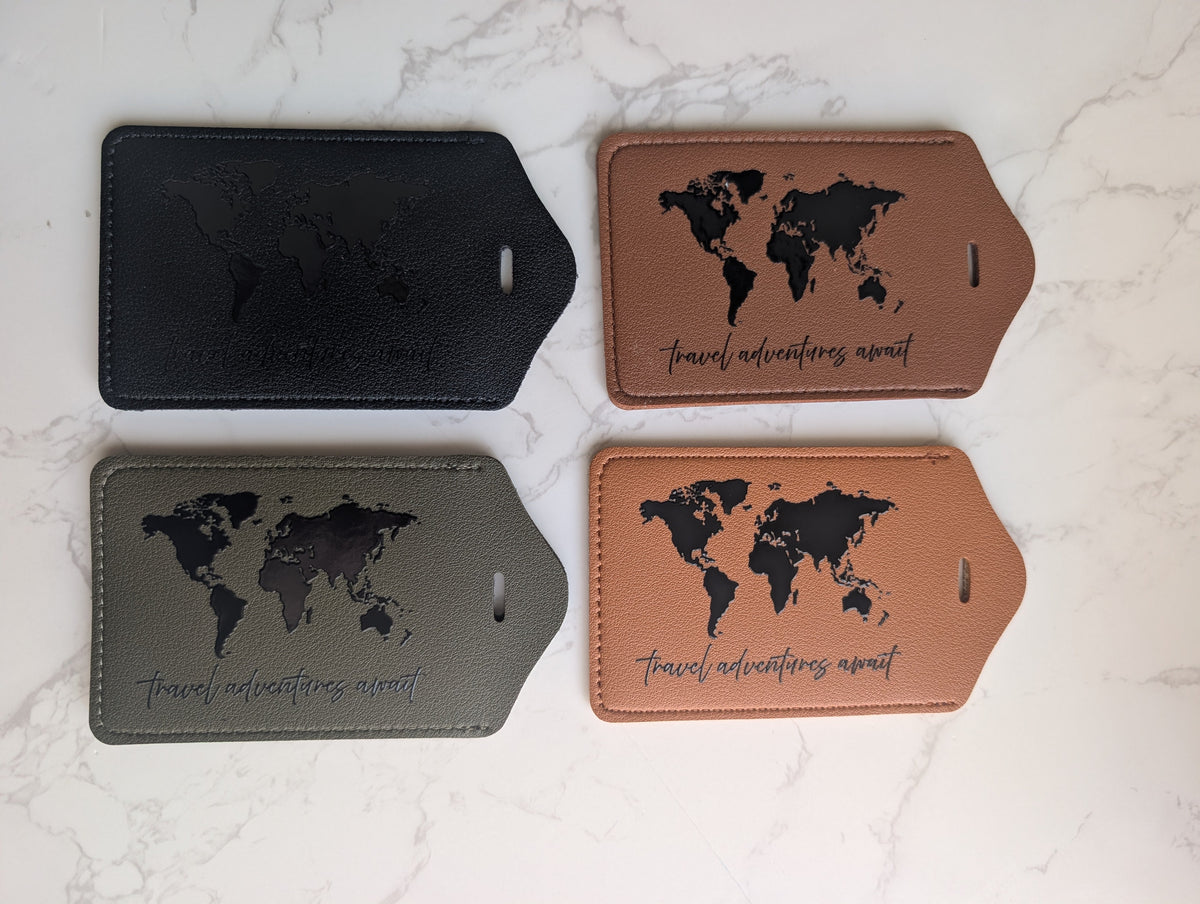 Four rectangular leather luggage tags arranged in a 2x2 grid on a white marble surface. The tags come in three colors: tan, olive green, and black.