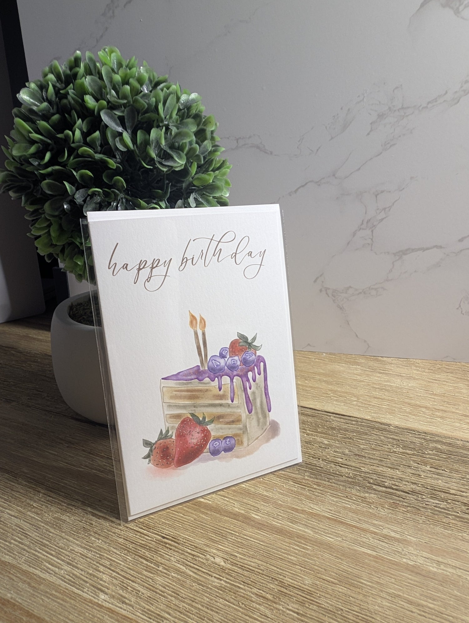 photo of watercolor notecard in portrait orientation, sitting flat on a wood table with a marble background. card features a watercolor illustration of a birthday hat and the message "Happy Birthday" in italic font