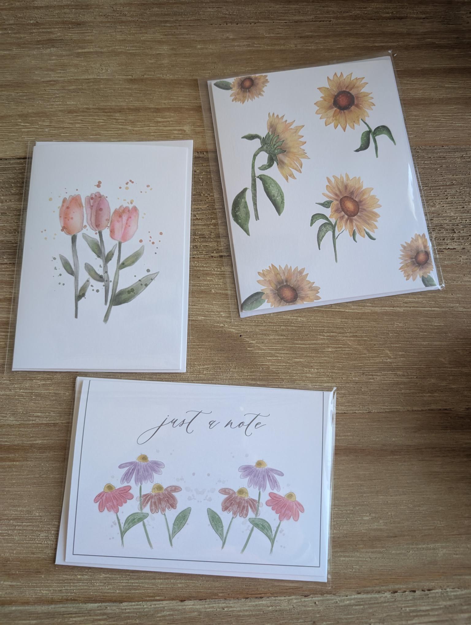 photo of three watercolor notecards n, sitting flat on a wood table. cards feature a watercolor illustration of: sunflower, tulips of daisy bundles