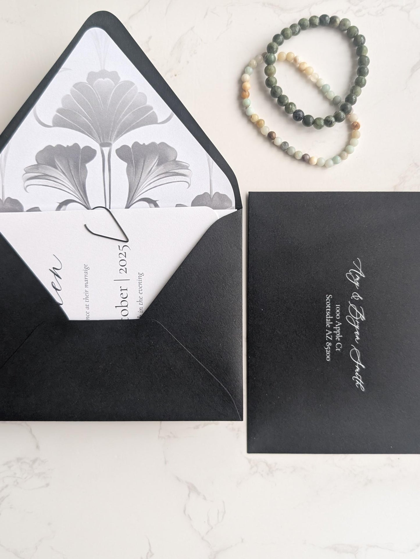 Black envelope with white interior featuring a floral design, next to a black card with white text on a light gray background.