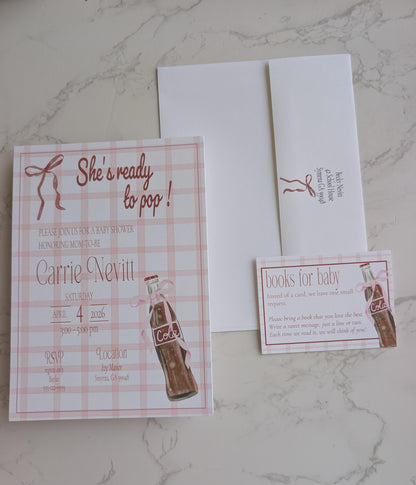 A baby shower invitation suite featuring a pink and white gingham card with 'She's ready to pop!' text, a white envelope, and a matching 'books for baby' RSVP card, laid out on a light marble surface.