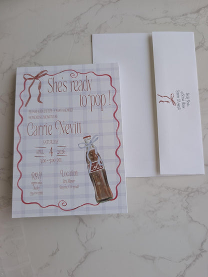 A printed invitation card with a vintage-style red script border and a bottle illustration, placed on a white envelope on a light marble surface. The invitation reads 'She's ready to pop!' and 'Carrie & Nevitt'.