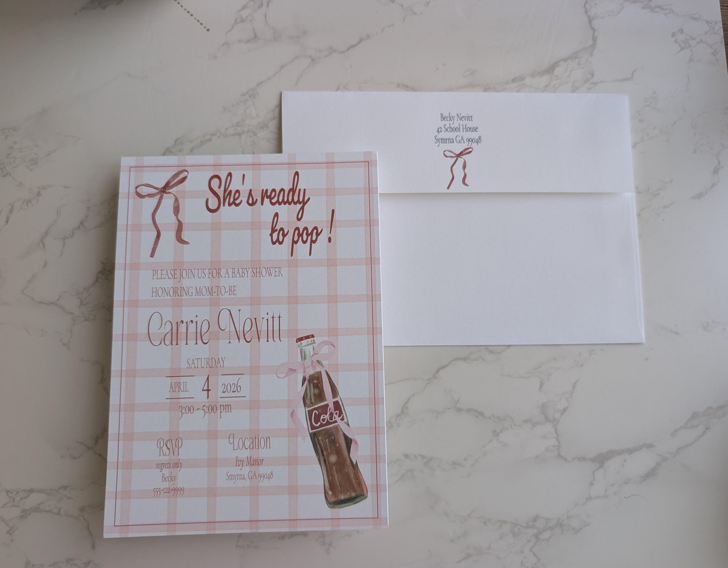 A square bridal shower invitation card with a pink plaid pattern and a vintage-style cola bottle illustration, placed next to a white envelope on a light marble surface.