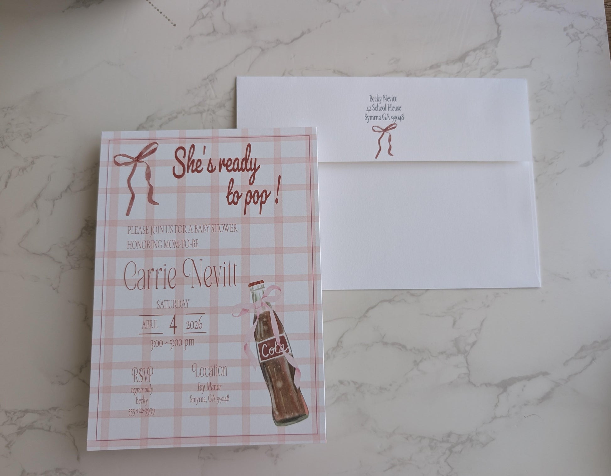 A square bridal shower invitation card with a pink plaid pattern and a vintage-style cola bottle illustration, placed next to a white envelope on a light marble surface.