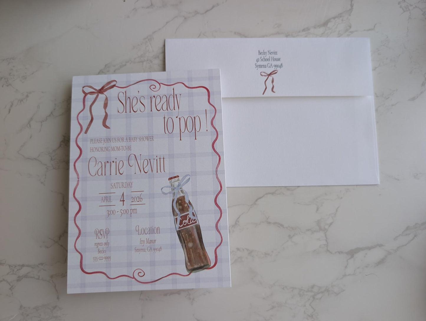 A personalized baby shower invitation card featuring a vintage Coca-Cola bottle illustration and red script text on a white plaid background, shown next to a matching white envelope on a light marble surface.