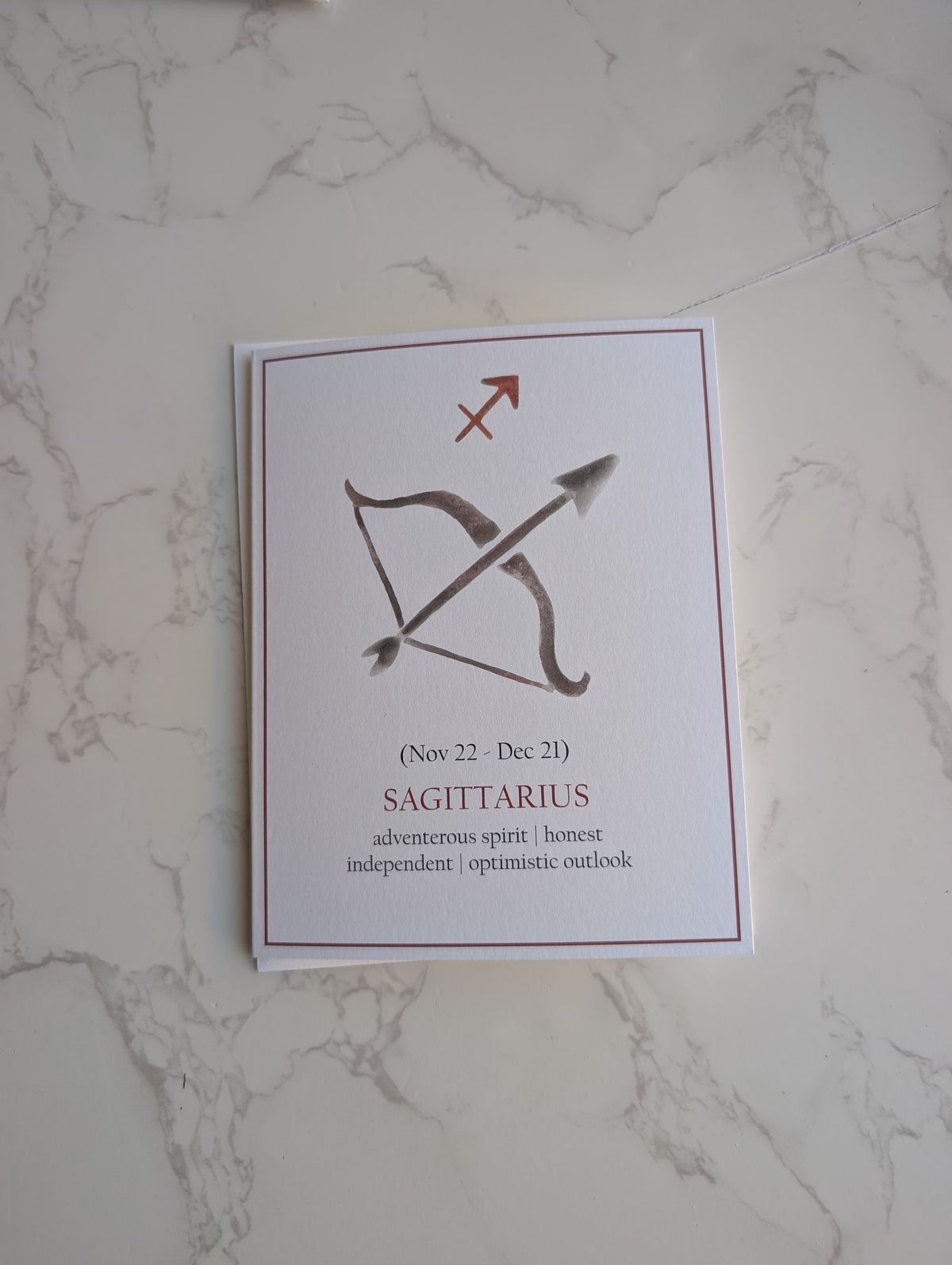 A square white card featuring the Sagittarius zodiac sign with a bow and arrow graphic, placed on a light marble surface.