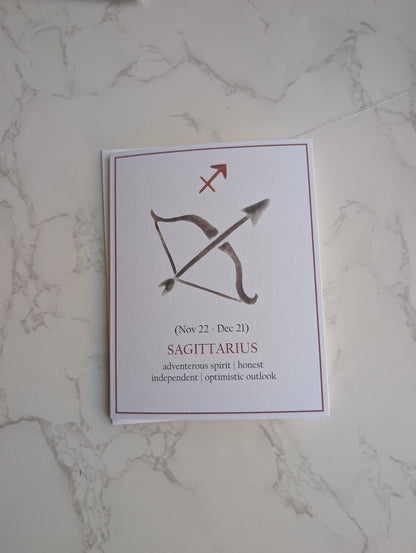 A square white card featuring the Sagittarius zodiac sign with a bow and arrow graphic, placed on a light marble surface.