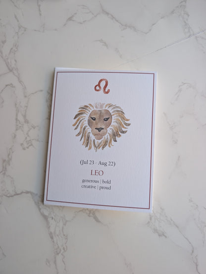 A white card featuring a watercolor lion illustration and the Leo zodiac sign, placed on a light marble surface.