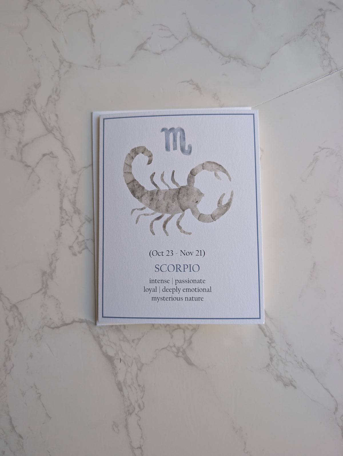 A vertical white card featuring the Scorpio zodiac sign with a gray watercolor scorpion illustration, placed on a white marble surface.