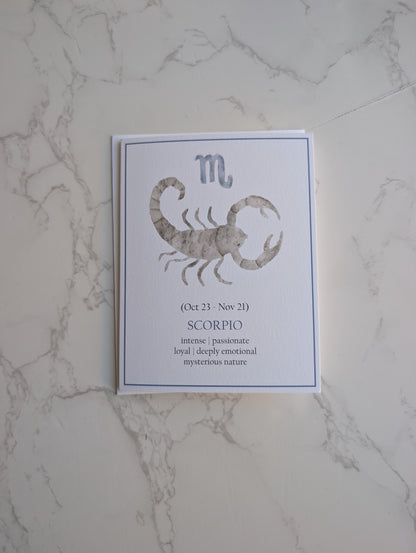 A vertical white card featuring the Scorpio zodiac sign with a gray watercolor scorpion illustration, placed on a white marble surface.