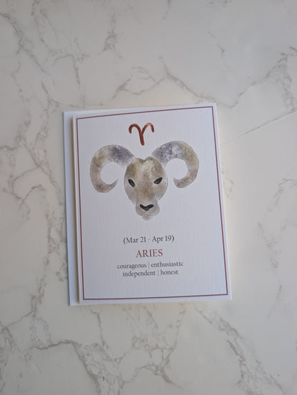 A rectangular greeting card featuring a watercolor ram illustration and the Aries zodiac sign, placed on a light marble surface.