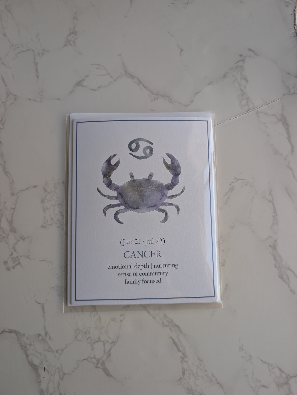 A square art print featuring a watercolor-style Cancer zodiac crab and astrological symbol, displayed on a light marble-patterned surface.