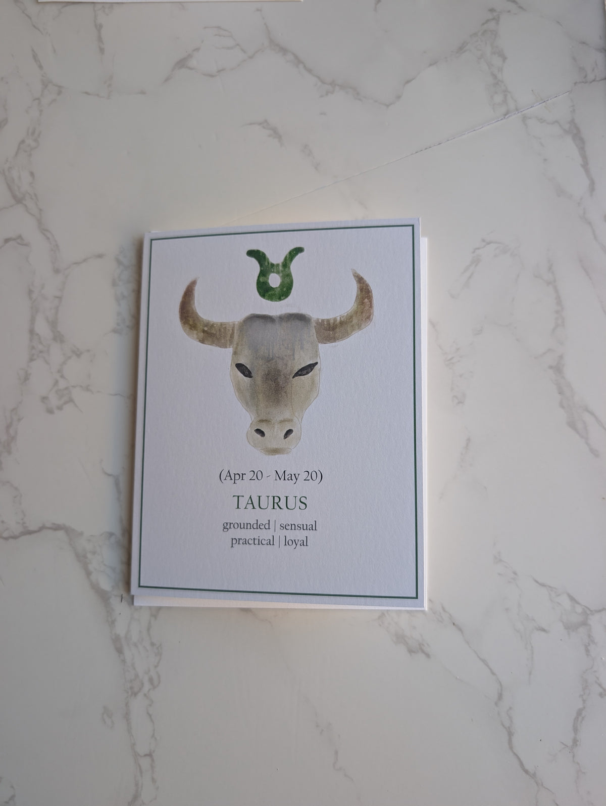 A zodiac-themed greeting card featuring a watercolor illustration of a Taurus bull head with a green horn, displayed on a white marble surface.