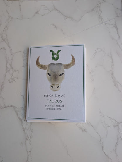 A zodiac-themed greeting card featuring a watercolor illustration of a Taurus bull head with a green horn, displayed on a white marble surface.