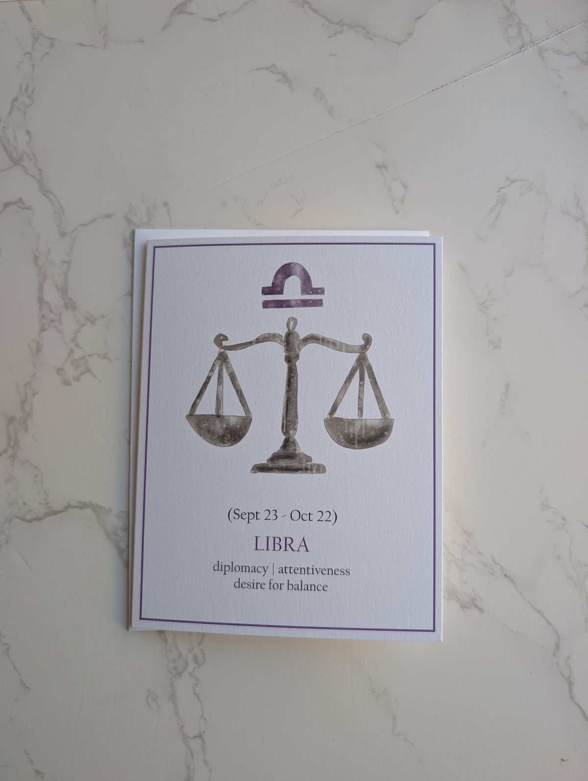 A square white card featuring a Libra zodiac design with a balance scale illustration and text, placed on a light marble surface.