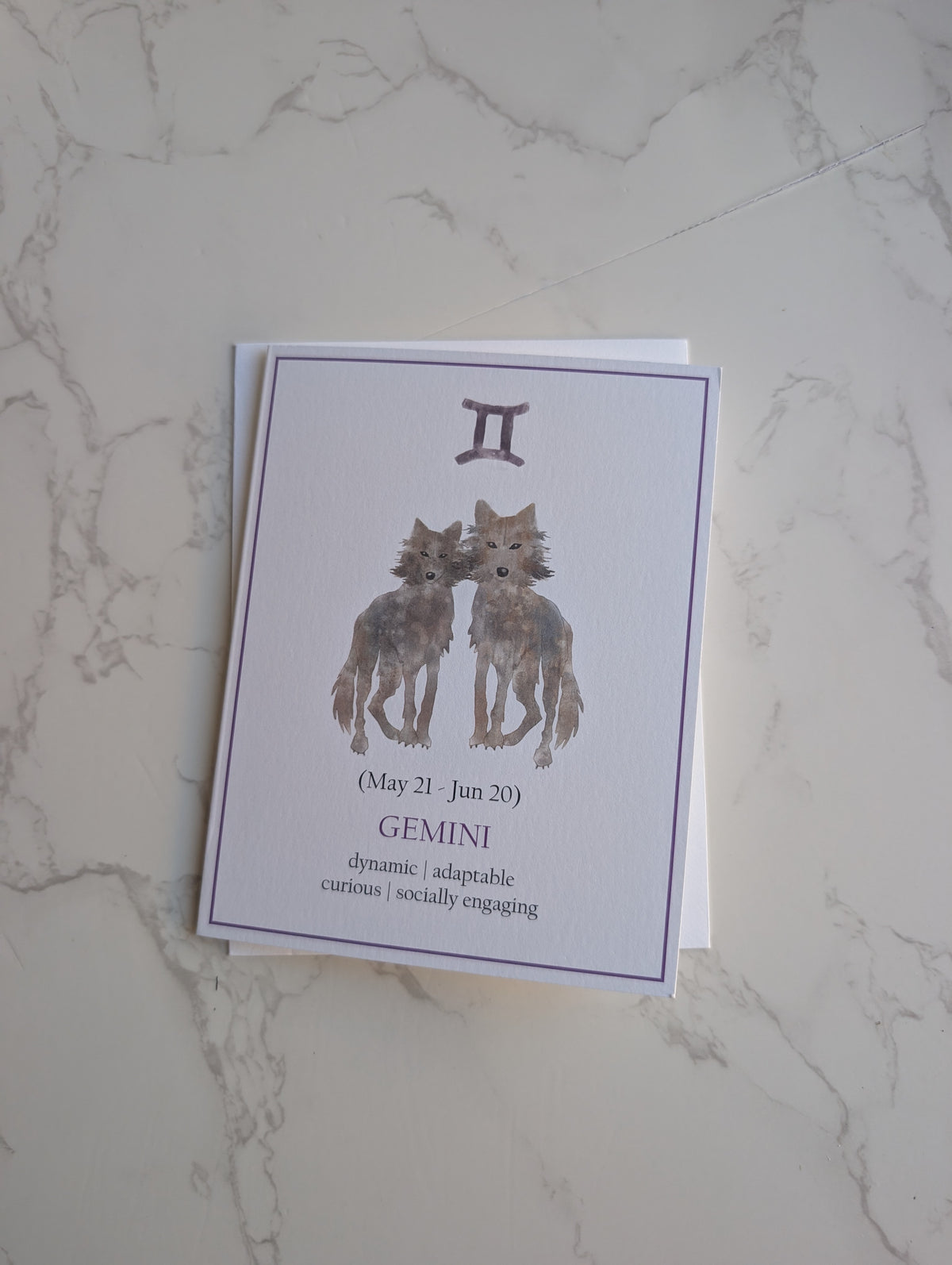 A square greeting card featuring a Gemini zodiac design with two illustrated wolves and astrological text, placed on a white marble surface.
