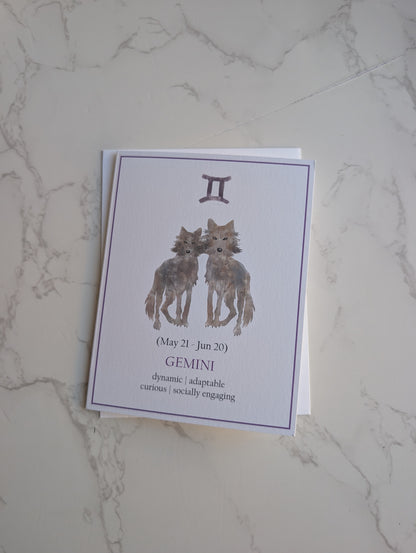 A square greeting card featuring a Gemini zodiac design with two illustrated wolves and astrological text, placed on a white marble surface.