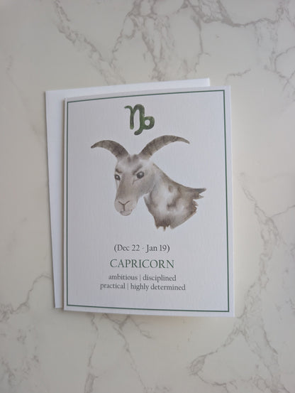 A zodiac-themed card featuring a watercolor illustration of a Capricorn goat head with the letter 'C' above it, placed on a white envelope against a light marble background.
