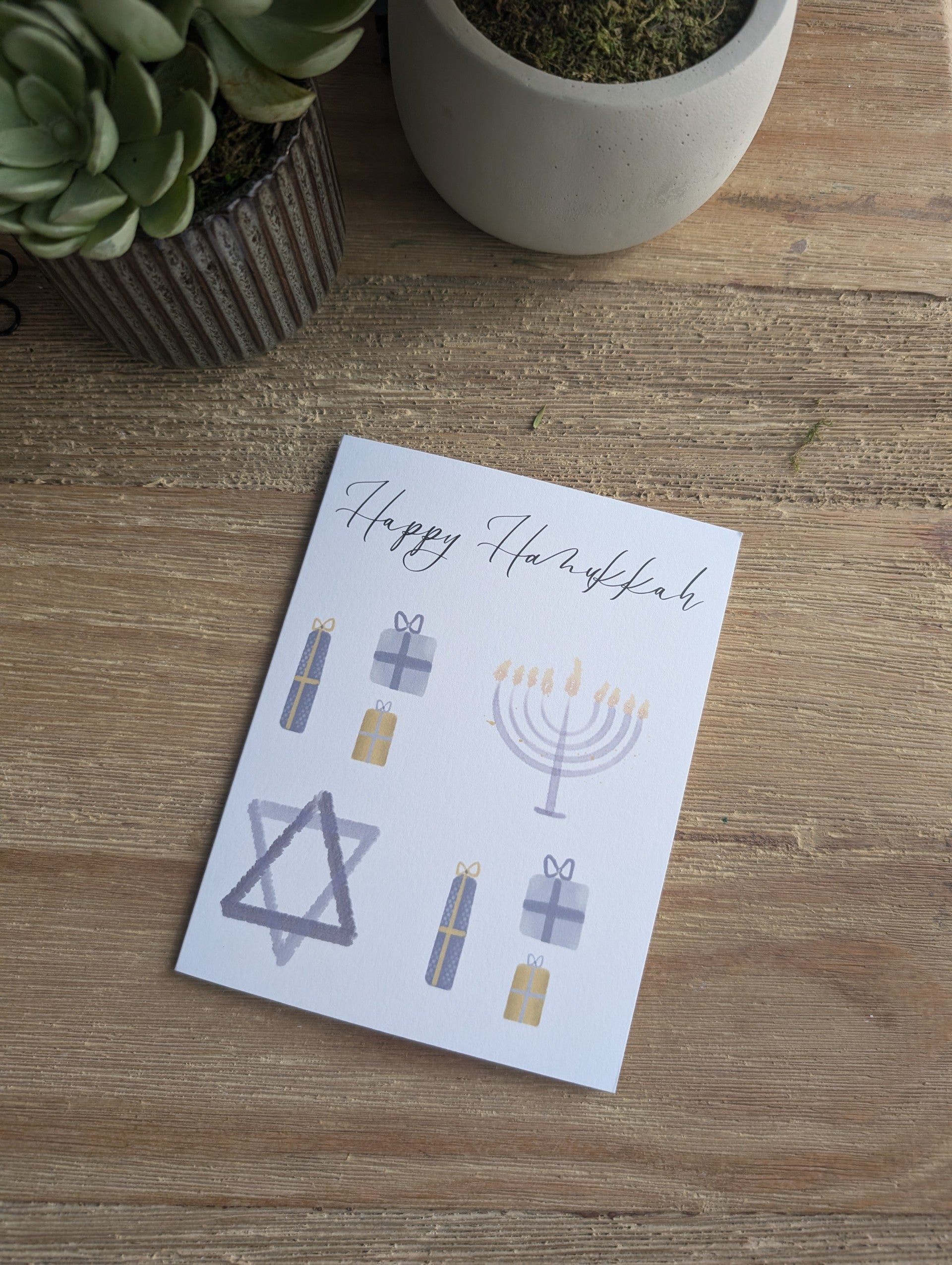 A folded notecard with a Hanukkah-themed watercolor design, including a Menorah and Star of David, accompanied by a white envelope, placed on a wooden surface.