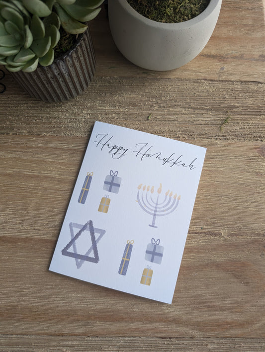 A folded notecard with a Hanukkah-themed watercolor design, including a Menorah and Star of David, accompanied by a white envelope, placed on a wooden surface.