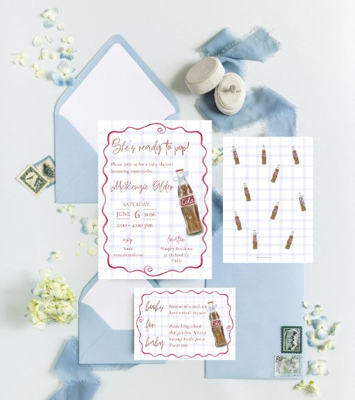 A styled flat-lay of a baby shower invitation suite featuring a main invitation card, an envelope, a shower checklist card, and a shower game card, all decorated with a vintage cola bottle illustration and pink borders, arranged on a light blue fabric background with baby shoes and flowers.