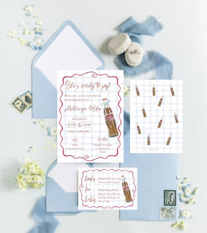 A styled flat-lay of a baby shower invitation suite featuring a main invitation card, an envelope, a shower checklist card, and a shower game card, all decorated with a vintage cola bottle illustration and pink borders, arranged on a light blue fabric background with baby shoes and flowers.