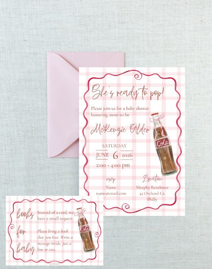 A baby shower invitation card with a pink gingham border and a retro cola bottle illustration, shown with a matching pink envelope on a light textured fabric background. A smaller matching card is placed in the lower left corner.