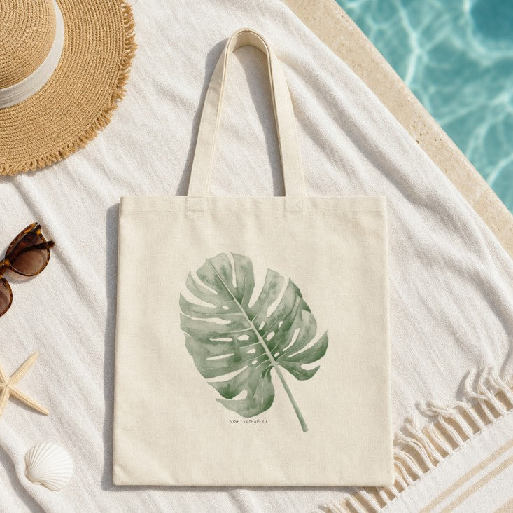 A cream canvas tote bag with a green monstera leaf print, styled on a white beach towel next to a straw hat, sunglasses, a starfish, and a seashell near a swimming pool.