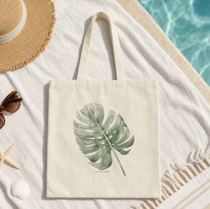 A cream canvas tote bag with a green monstera leaf print, styled on a white beach towel next to a straw hat, sunglasses, a starfish, and a seashell near a swimming pool.