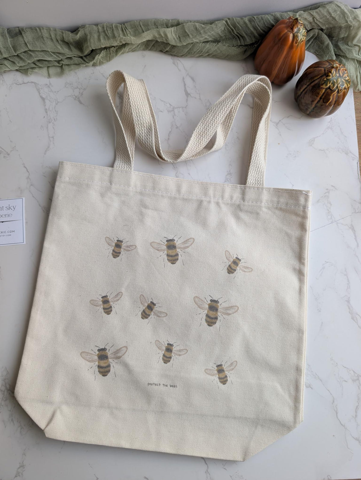 An off-white canvas tote bag with printed illustrations of bees, laid flat on a white marble surface with decorative props including a green fabric runner and two small pumpkins.