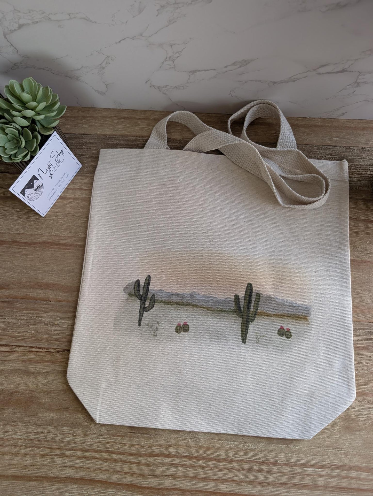 Tote bag with a desert landscape watercolor design featuring cacti on a wooden surface.