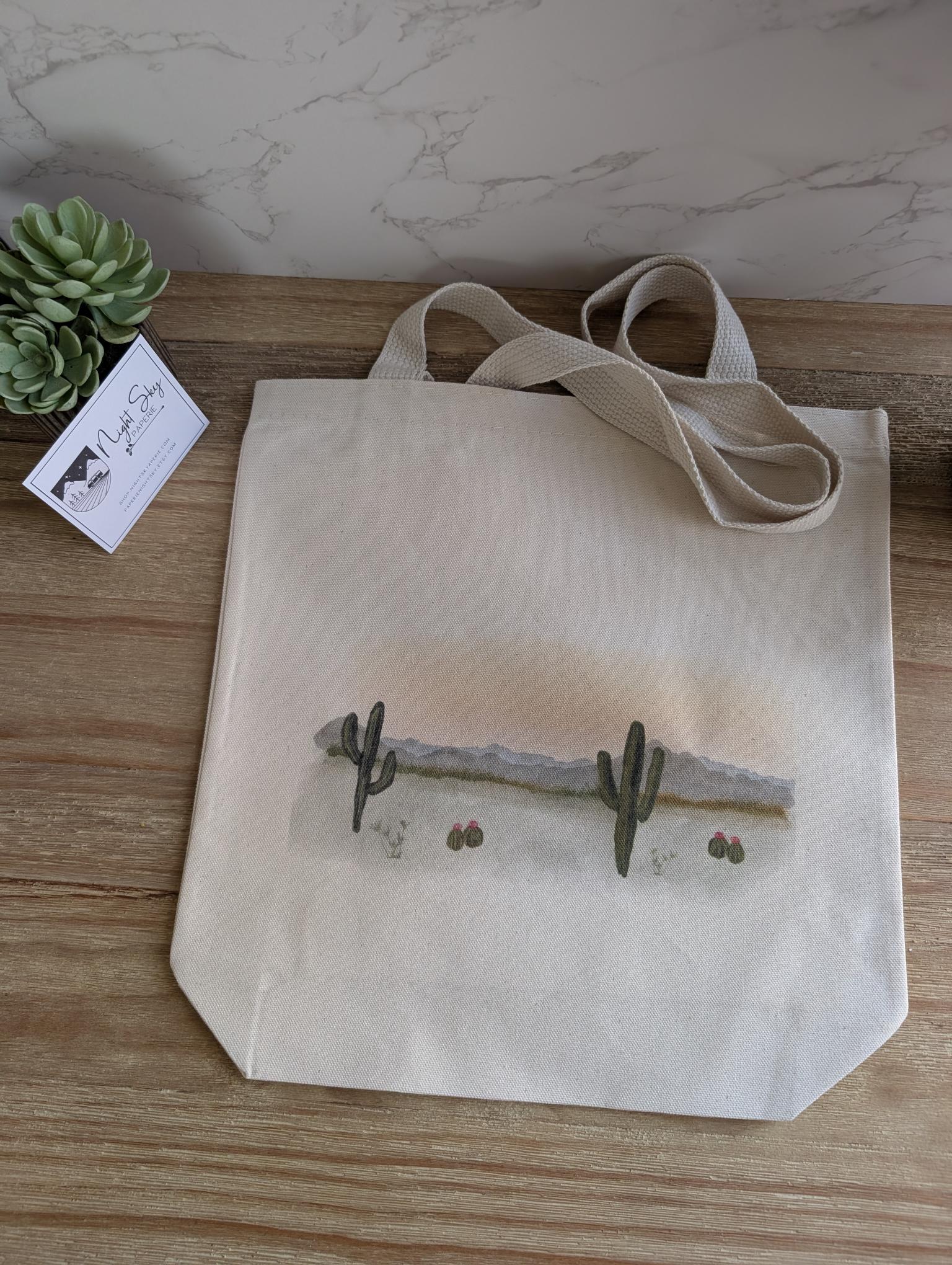Tote bag with a desert landscape watercolor design featuring cacti on a wooden surface.