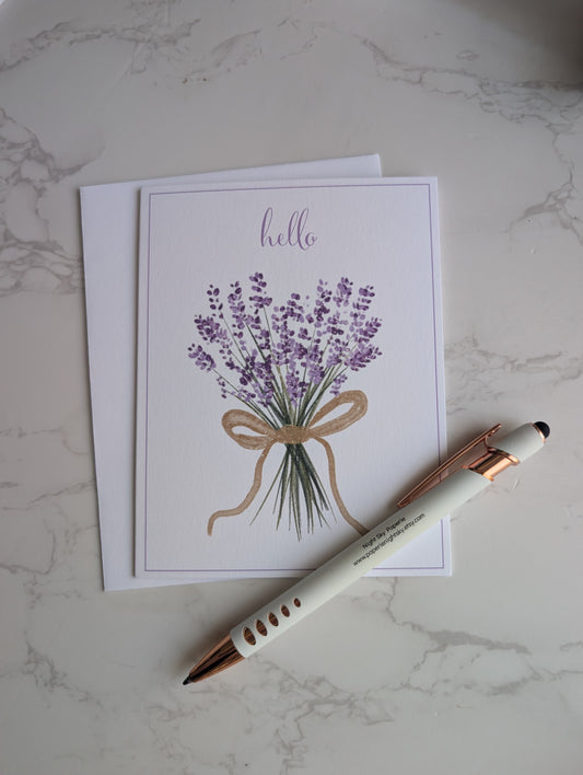 A square greeting card with a watercolor illustration of a lavender bouquet and the word 'hello' at the top, placed on a white envelope over a light marble surface with a white and rose gold pen resting diagonally across the lower right.