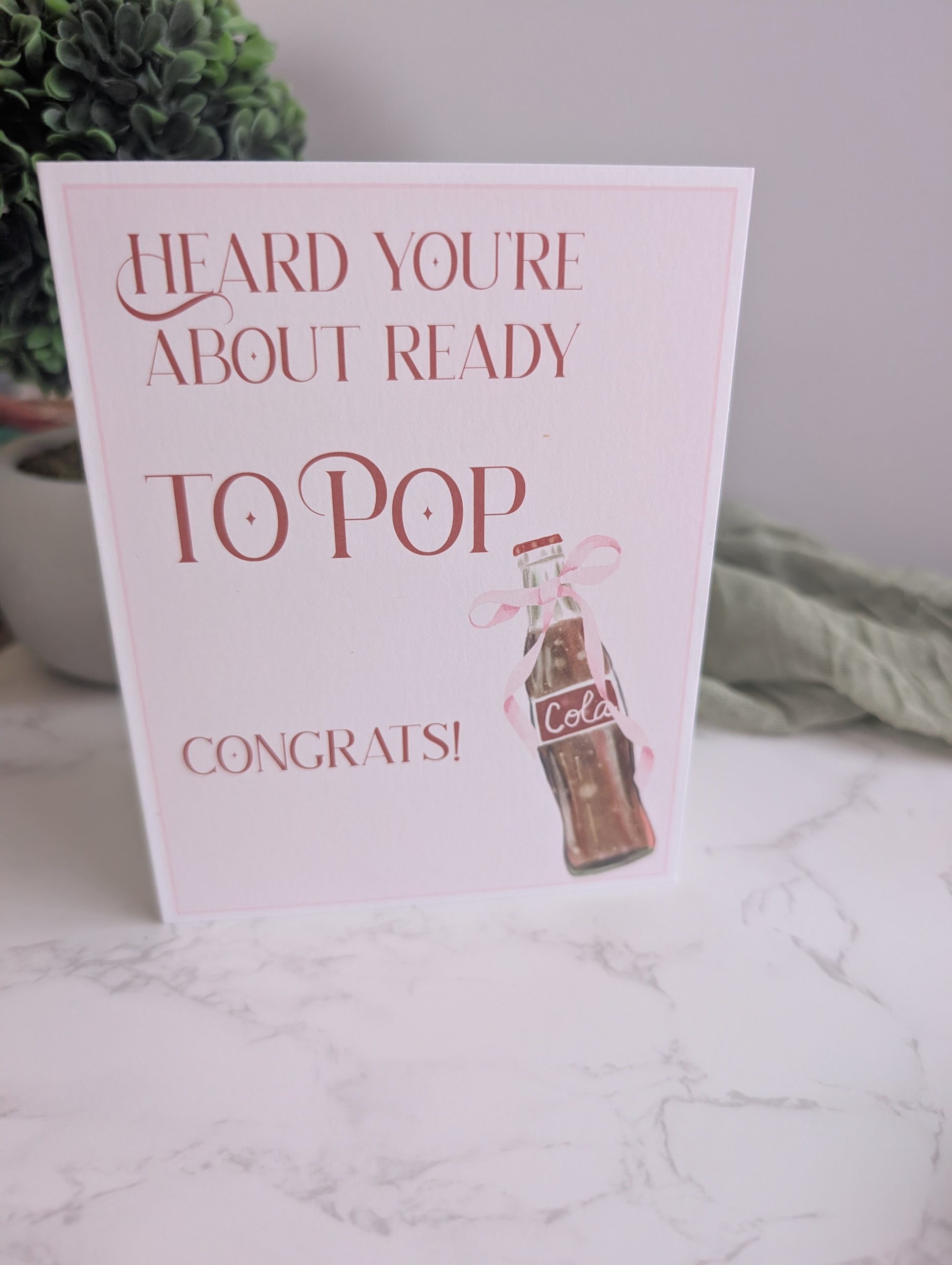 Greeting card with ' Heard you're about ready to pop. Congrats!' text and a bottle illustration on a marble surface.