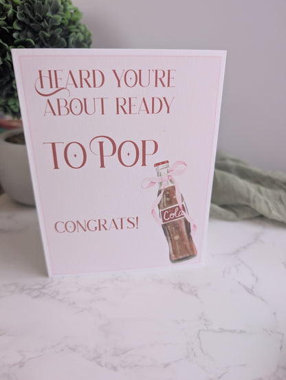 Greeting card with ' Heard you're about ready to pop. Congrats!' text and a bottle illustration on a marble surface.