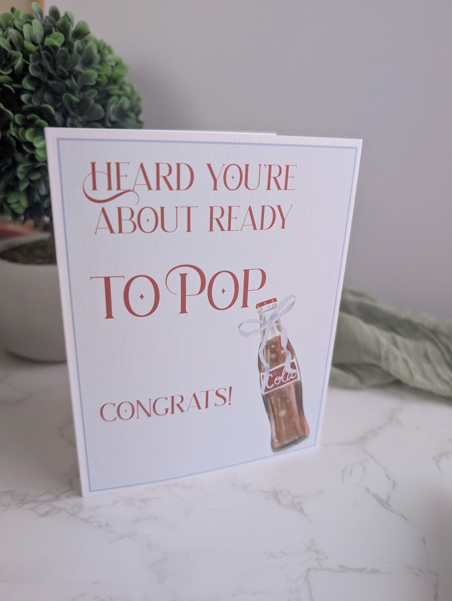Greeting card with text 'Heard you're about ready to pop congrats!' on a marble surface with a plant in the background.
