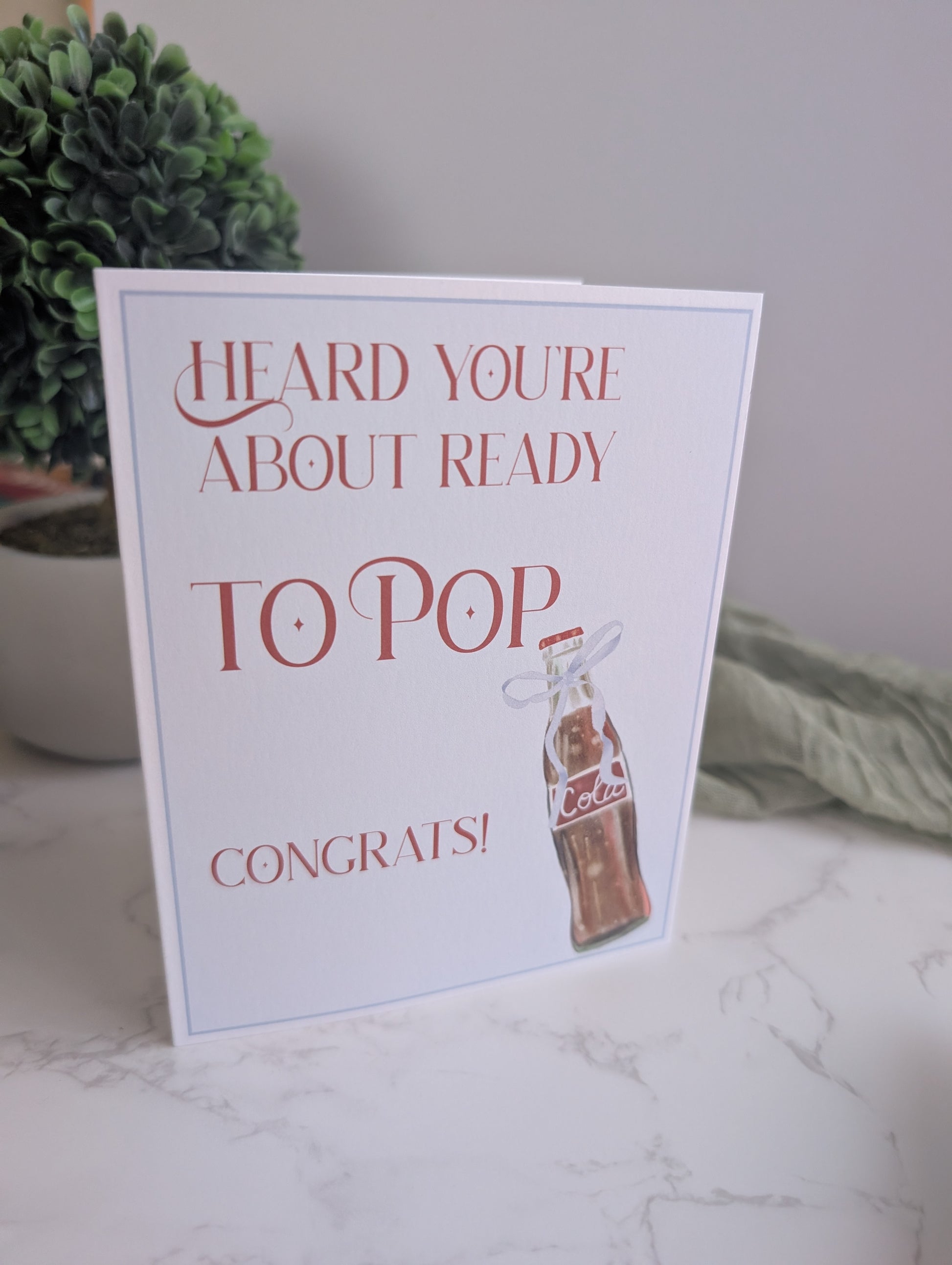Greeting card with text 'Heard you're about ready to pop congrats!' on a marble surface with a plant in the background.