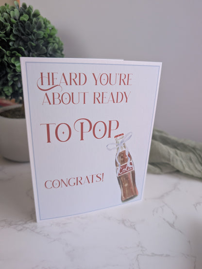Greeting card with text 'Heard you're about ready to pop congrats!' on a marble surface with a plant in the background.