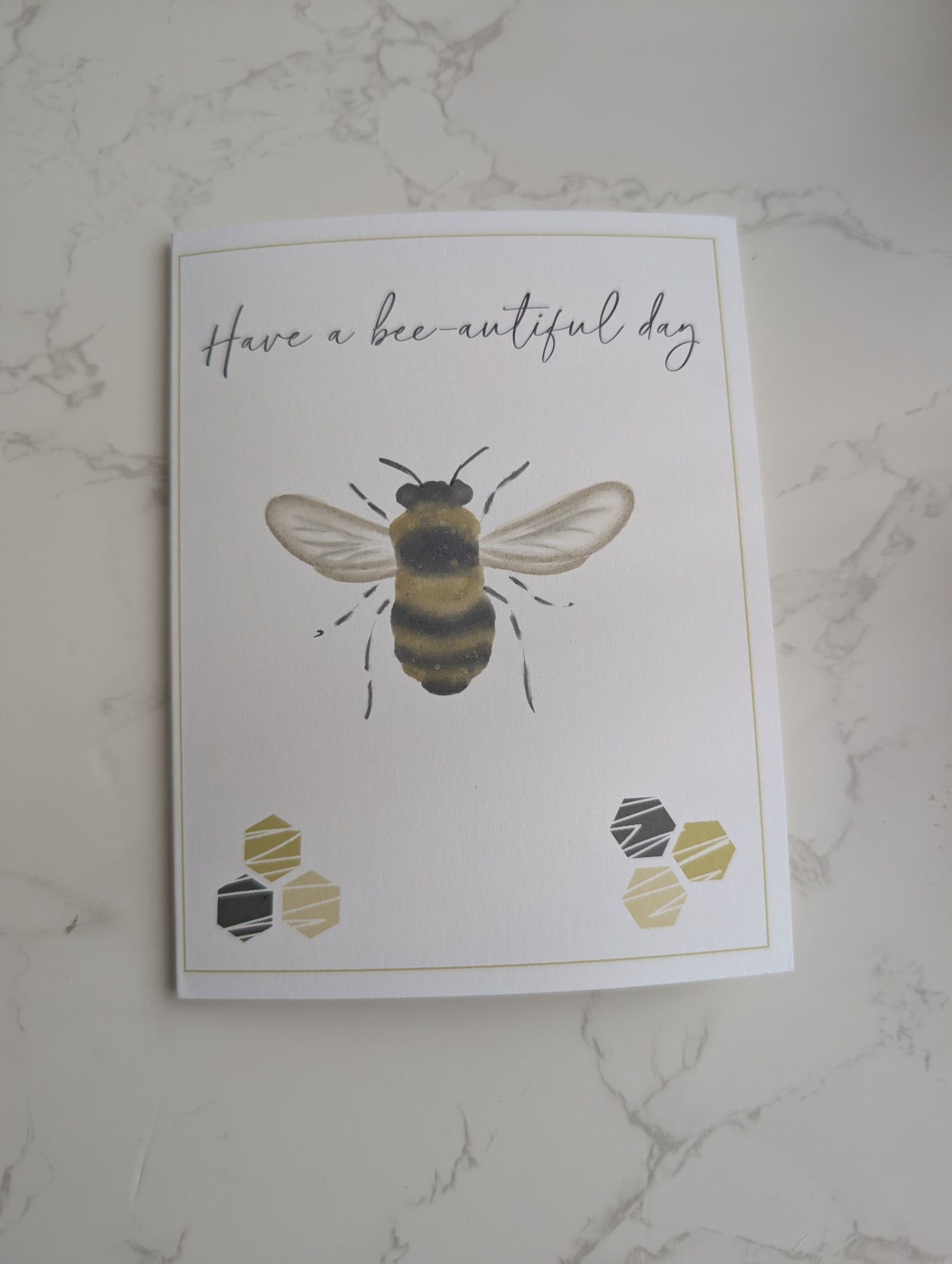 Greeting card with a bee illustration and 'Have a bee-autiful day' text on a marble surface.