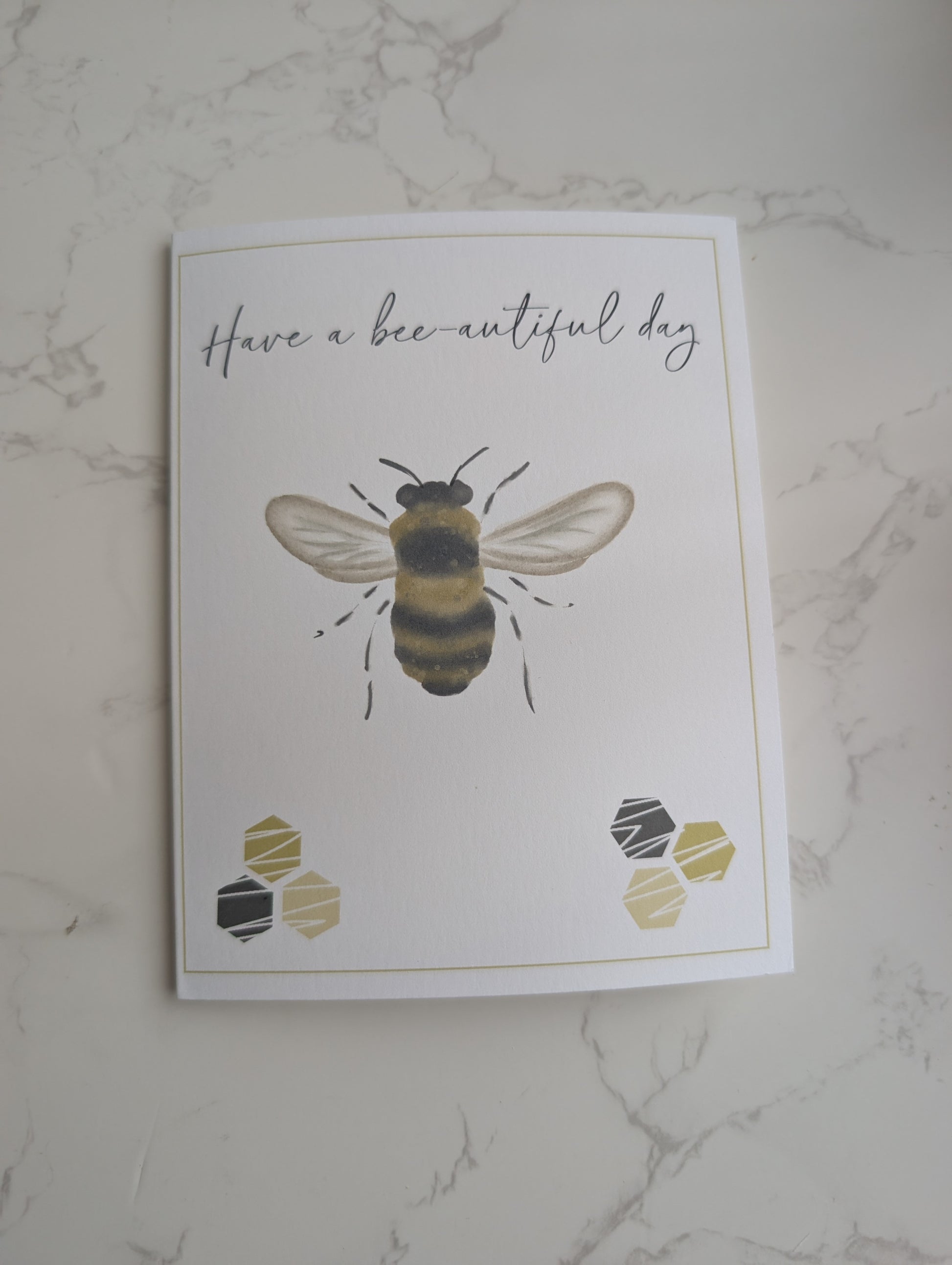 Greeting card with a bee illustration and 'Have a bee-autiful day' text on a marble surface.
