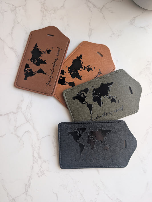 Four leather luggage tags in brown, tan, gray, and black, each featuring an embossed world map and the phrase 'true adventure does not go alone' on a white marble background.