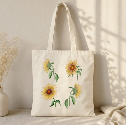 An off-white canvas tote bag with four watercolor-style sunflower prints, displayed on a neutral surface with soft shadows and dried flowers in a vase to the side.