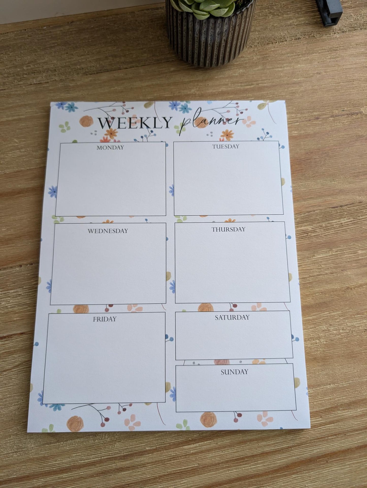Weekly planner notepad with watercolor embellishments, displayed on a wooden surface with a potted plant in the background.