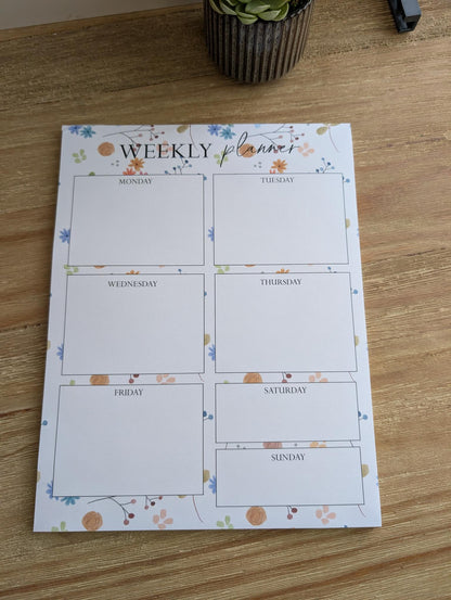 Weekly planner notepad with watercolor embellishments, displayed on a wooden surface with a potted plant in the background.